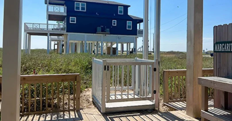 Affordable Elevator Website – Homepage Lifestyle hero Elevated beach house with stilts and spacious decks, home elevator in foreground for easy access, surrounded by lush grass and blue sky.