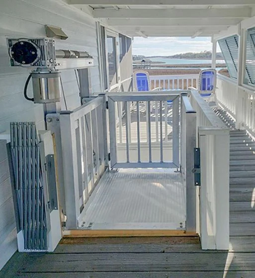 Outdoor home elevator with white railing, leading to a deck with blue chairs, overlooking a waterfront view.