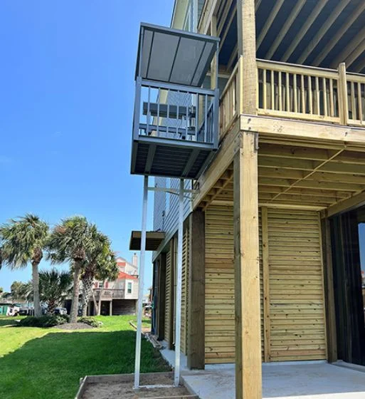 Outdoor home elevator on a wooden porch of a raised beach house, palm trees and neighboring homes in the background.