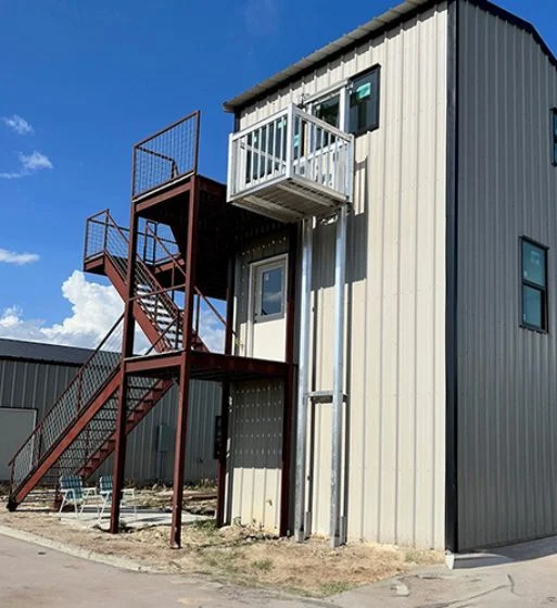 Exterior view of a beige building featuring a cargo elevator with a metal platform and rails, connected to a second-story platform.