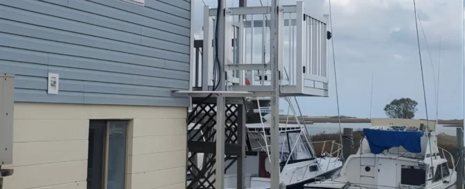 Outdoor home elevator next to a blue house by a marina, boats docked nearby under a cloudy sky.