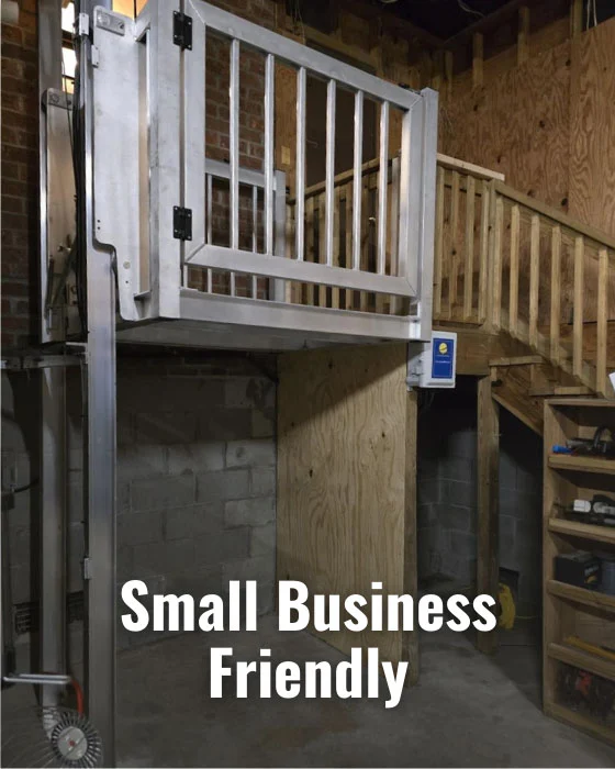 Metal platform lift labeled "Small Business Friendly," set in a workshop. Ideal for small spaces and home elevators, adjacent to wooden stairs.