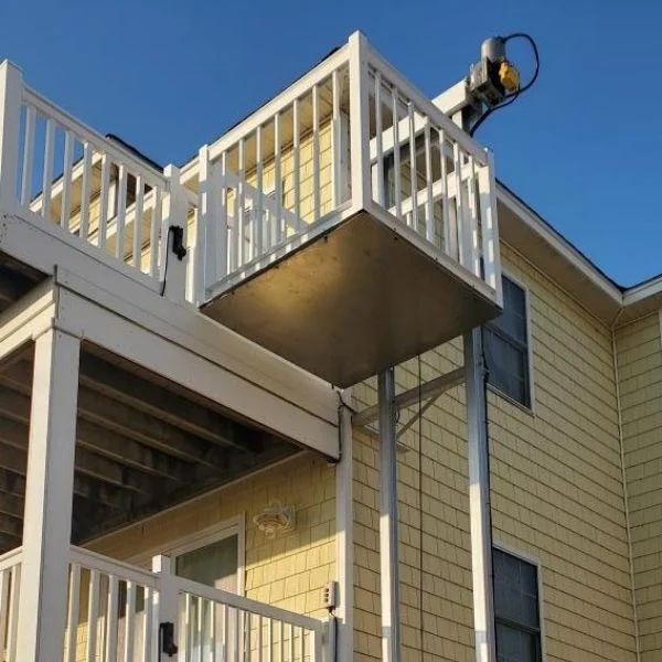 Outdoor Lift Outdoor residential platform with railing, used as a home elevator on a two-story yellow house, under a clear blue sky.