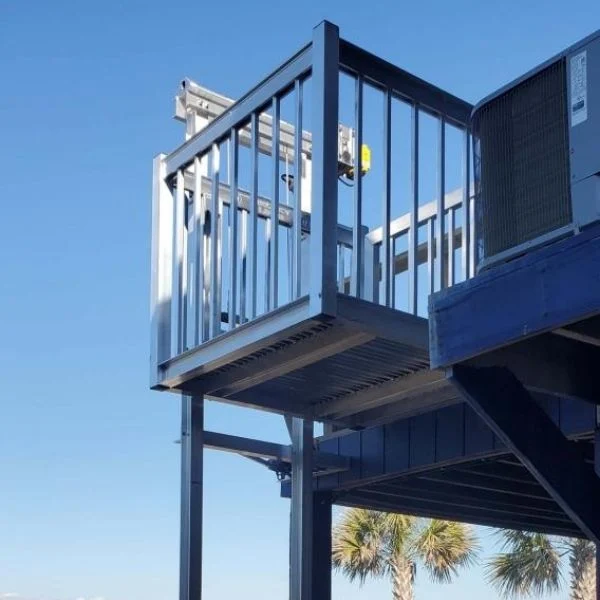 Outdoor Lift Outdoor home elevator platform with metal railings, partially shaded, against a clear blue sky and a glimpse of palm trees.