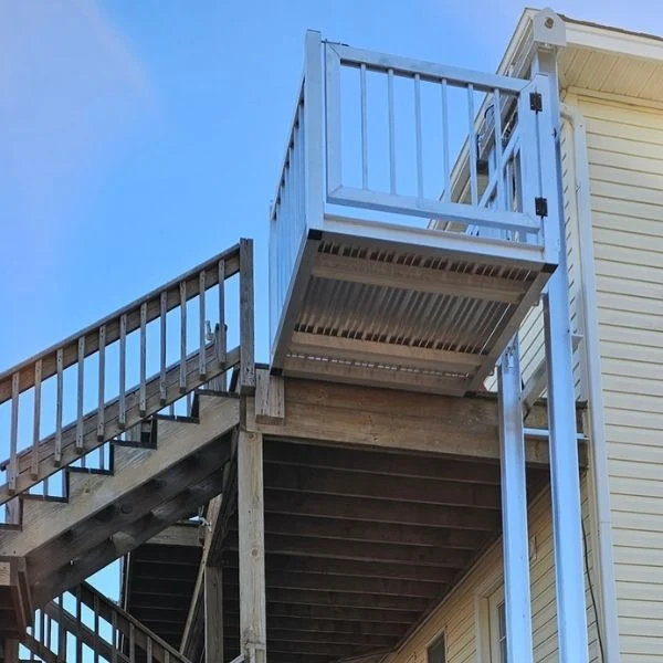 Outdoor home elevator with a metal platform next to wooden stairs, attached to a yellow house.
