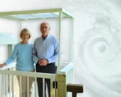 Elderly couple stands on home elevator platform, smiling against a swirling hurricane backdrop.